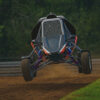 A 3D-rendered off-road buggy jumps mid-air on a dirt track, with its suspension fully extended and a blurred forest background resembling iRacing graphics.
