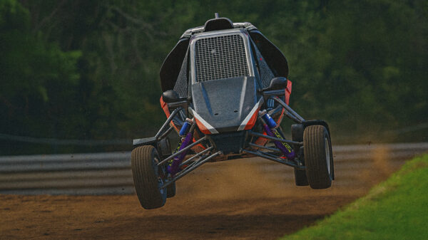 A 3D-rendered off-road buggy jumps mid-air on a dirt track, with its suspension fully extended and a blurred forest background resembling iRacing graphics.