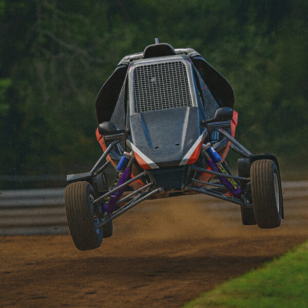 A 3D-rendered off-road buggy jumps mid-air on a dirt track, with its suspension fully extended and a blurred forest background resembling iRacing graphics.