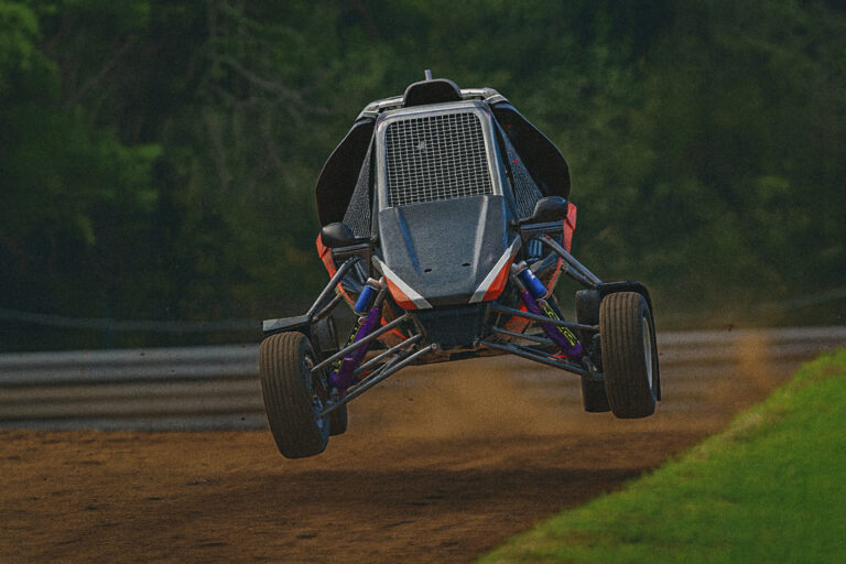 A 3D-rendered off-road buggy jumps mid-air on a dirt track, with its suspension fully extended and a blurred forest background resembling iRacing graphics.